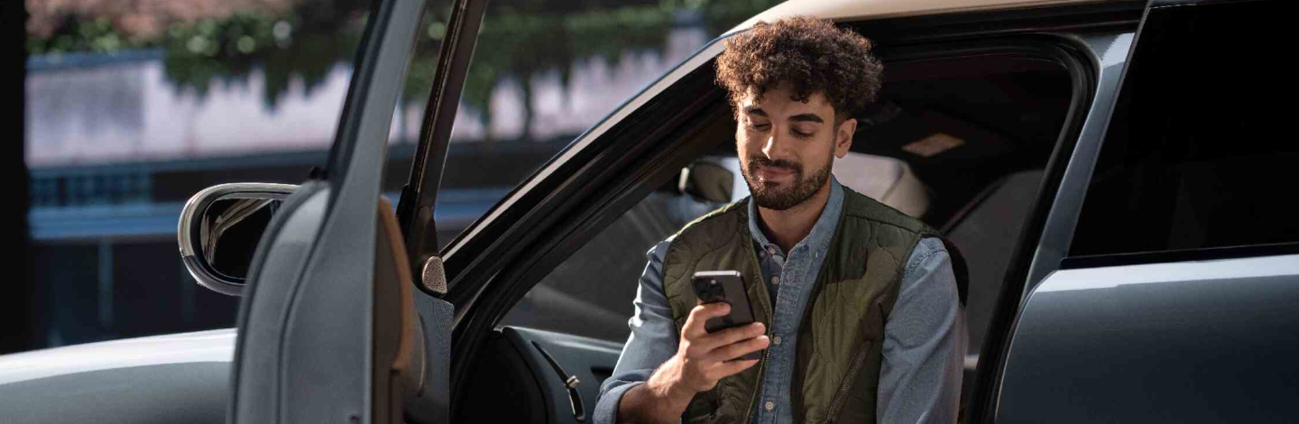 Mann sitzt in einem Auto, schaut auf sein Smartphone, Autotür ist geöffnet.