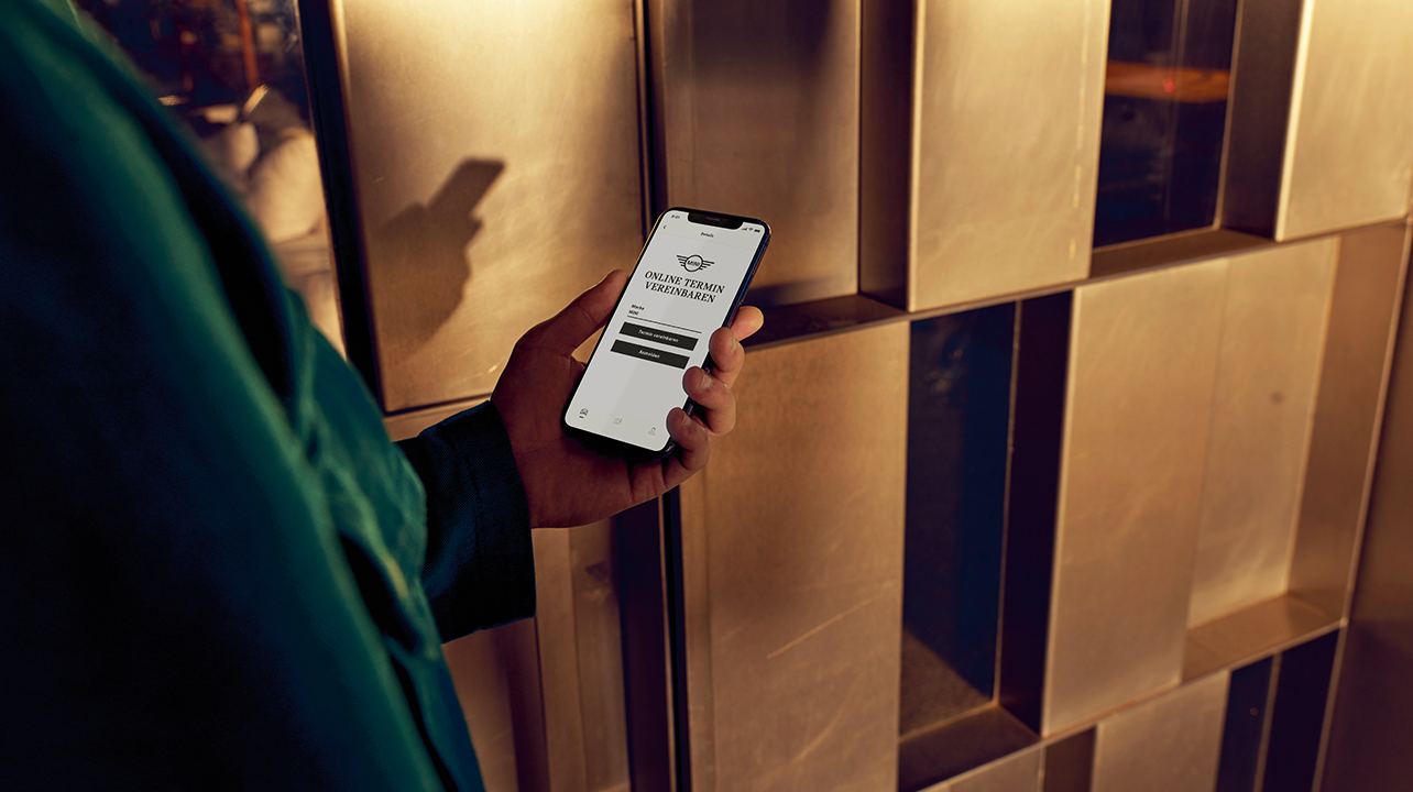 Person hält Smartphone mit Terminvereinbarung in der Hand, im Hintergrund metallene Wand.