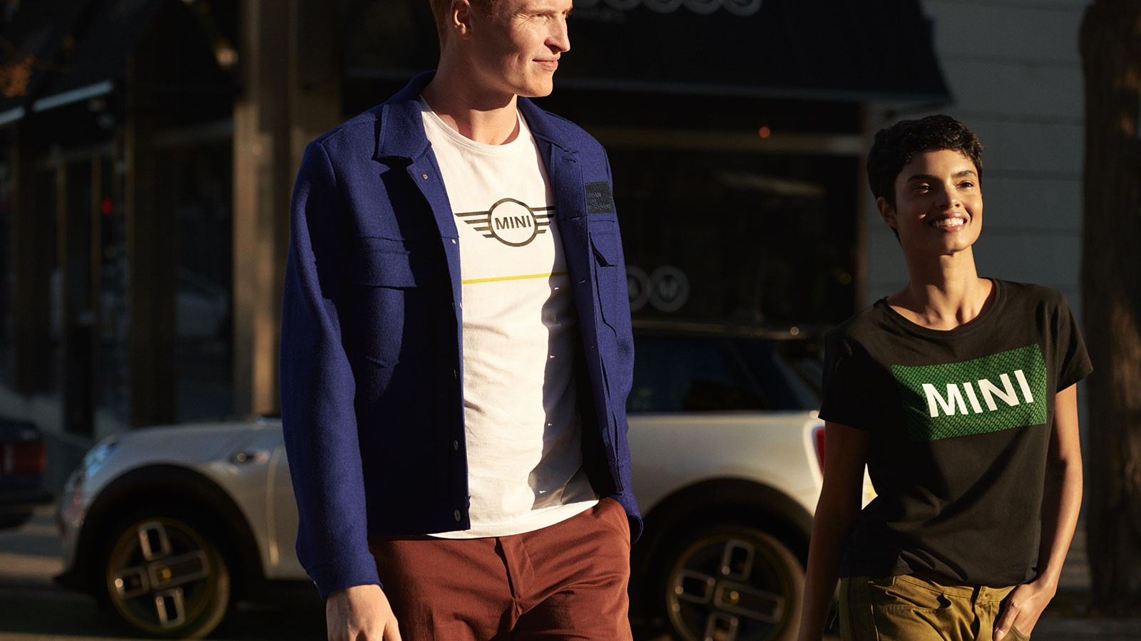 Zwei Personen in MINI-Shirts gehen vor einem geparkten weißen MINI, sonniger Tag.