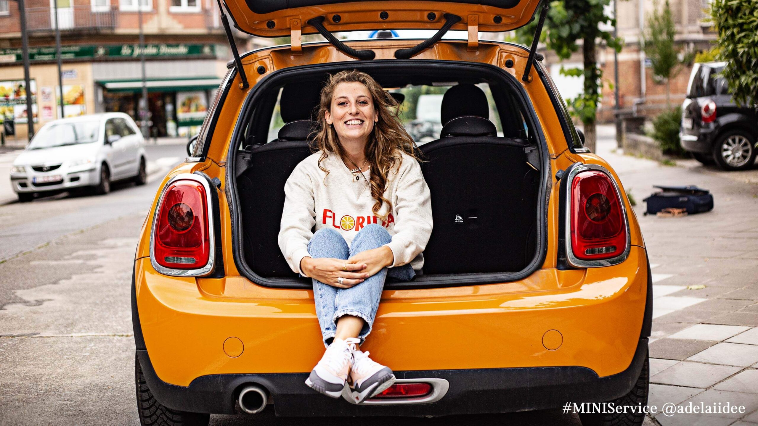 Frau sitzt lächelnd im geöffneten Kofferraum eines gelben MINI auf einer Straße.
