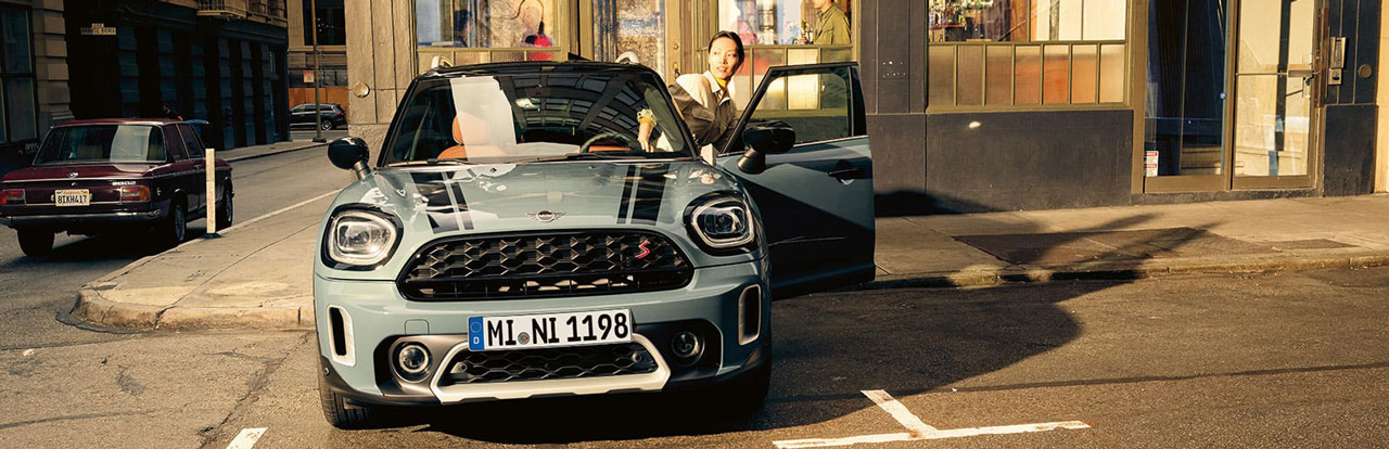 Grauer MINI Countryman auf Stadtstraße, Person steigt aus, Gebäude im Hintergrund.