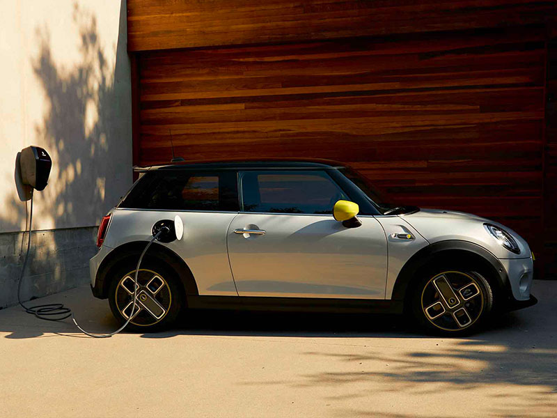 Silberner MINI Cooper SE wird an Ladestation geladen, steht vor Holzfassade.