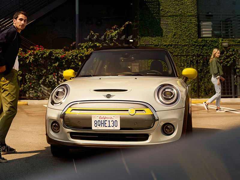 Silberner MINI Cooper SE mit gelben Akzenten, zwei Personen im Hintergrund auf einem Parkplatz.