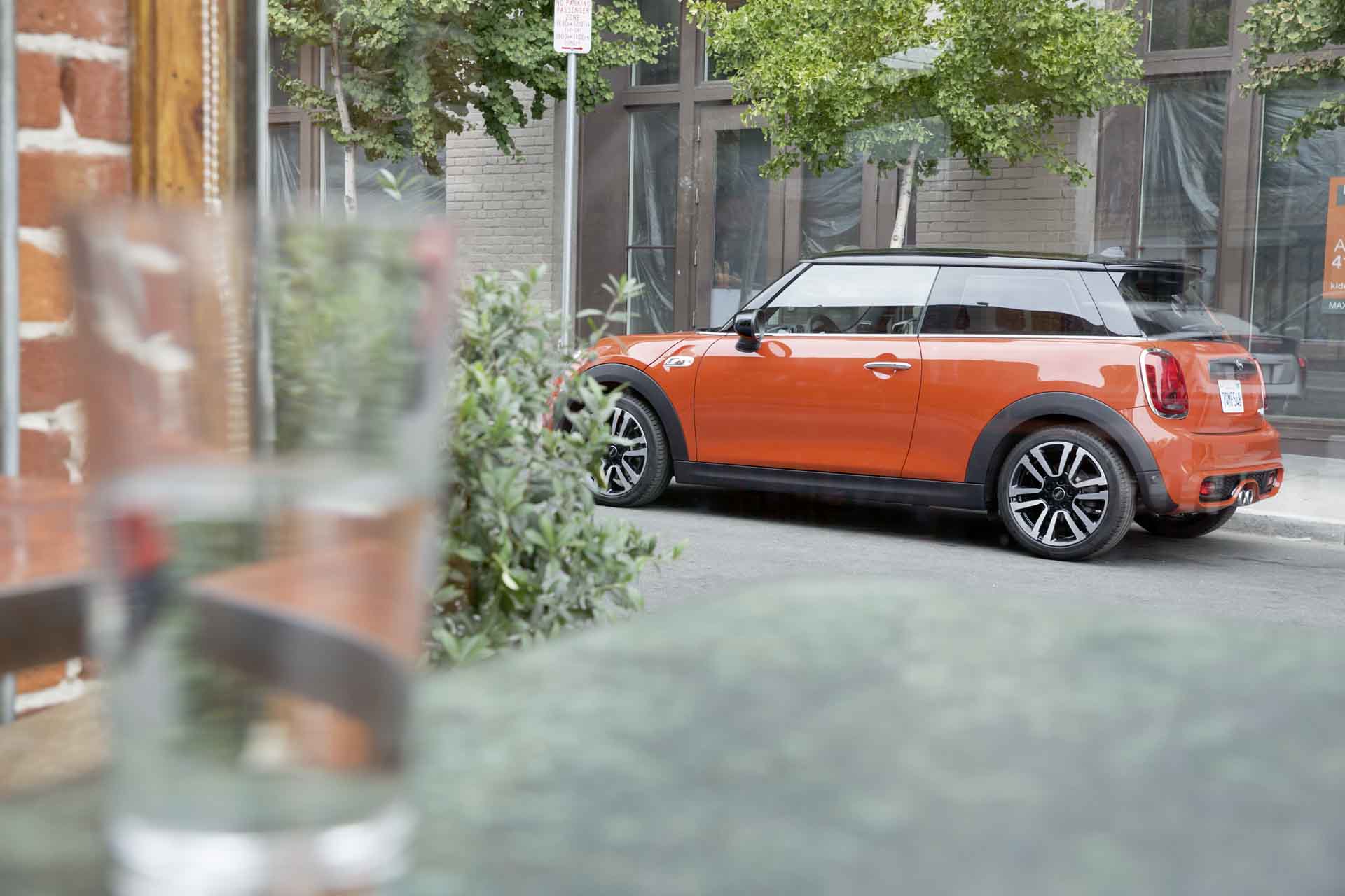 Ein oranger MINI Cooper steht am Straßenrand, Bäume und Gebäude im Hintergrund.