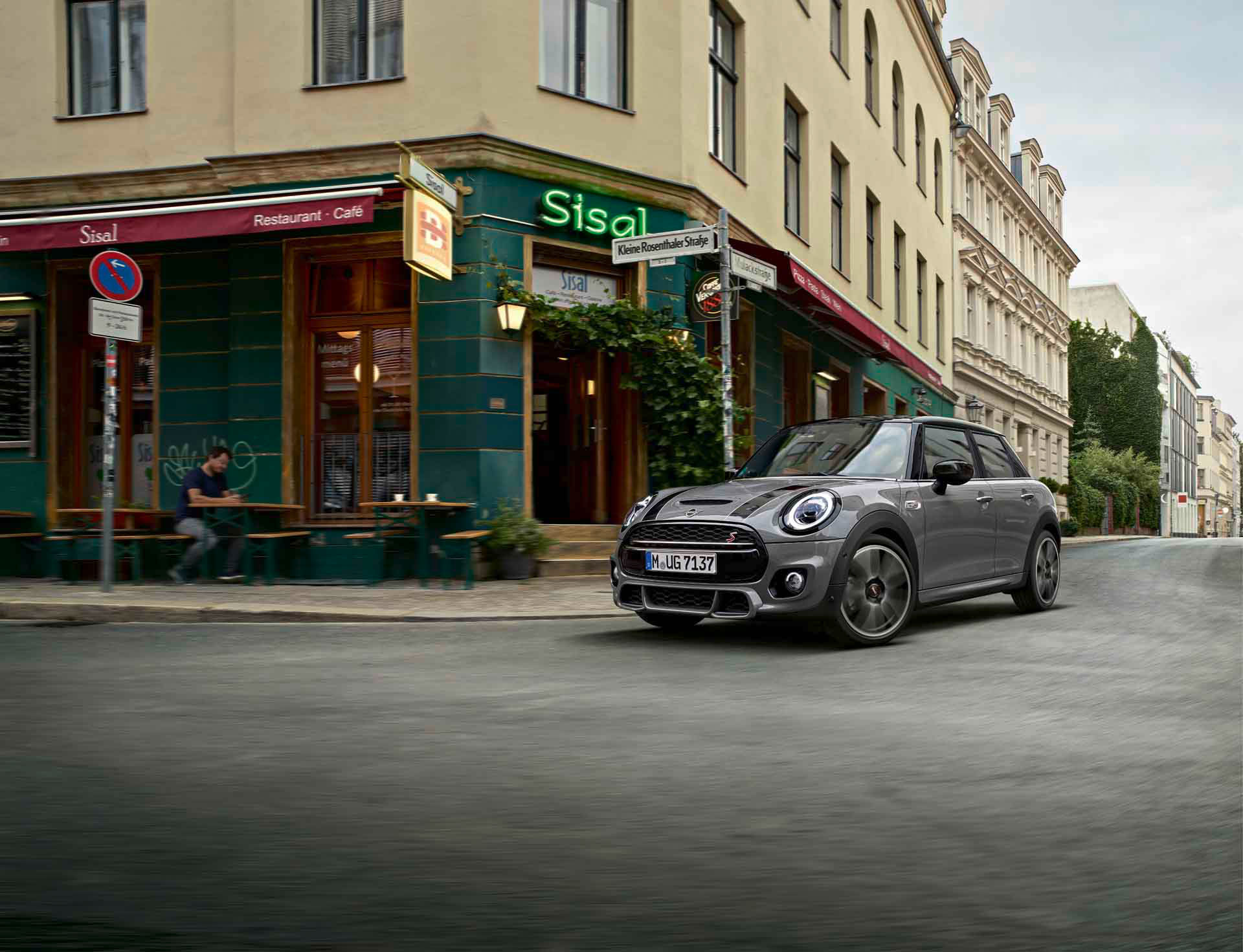 Grauer MINI Fünftürer parkt vor einem Eckcafé in einer städtischen Straßenszene.