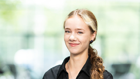 Eine lächelnde Frau mit blonden Haaren und schwarzer Bluse vor verschwommenem Hintergrund.