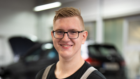 Ein junger Mann mit Brille lächelt in einer Autowerkstatt. Im Hintergrund verschwommene Fahrzeuge.