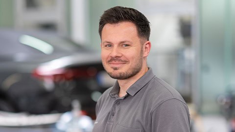 Ein Mann in grauem Hemd lächelt in einer Autowerkstatt, unscharfes Auto im Hintergrund.