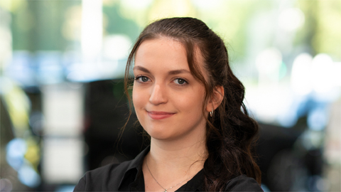 Eine Frau mit dunklem Haar lächelt in die Kamera, Hintergrund ist unscharf.