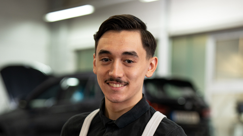 Ein Mann mit Schnurrbart lächelt in die Kamera, im Hintergrund unscharfe Autos.