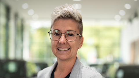 Eine Person mit Brille und kurzem Haar lächelt in einem hellen Raum.