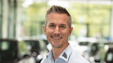 Ein Mann in einem hellblauen Hemd lächelt in die Kamera, unscharfe Autos im Hintergrund.
