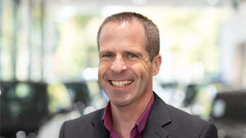 Ein lächelnder Mann in einem Anzug vor einem unscharfen Hintergrund in einem Autohaus.