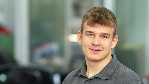 Junger Mann in grauem Hemd lächelt in die Kamera, unscharfer Hintergrund in einem Autohaus.