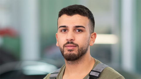 Ein Mann mit dunklen Haaren und Bart schaut in die Kamera, unscharfer Hintergrund.