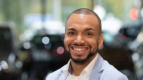 Ein Mann lächelt in die Kamera, trägt ein helles Sakko; verschwommener Hintergrund.