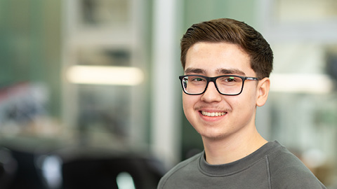 Ein junger Mann mit Brille lächelt in die Kamera, im Hintergrund ein unscharfes Auto.