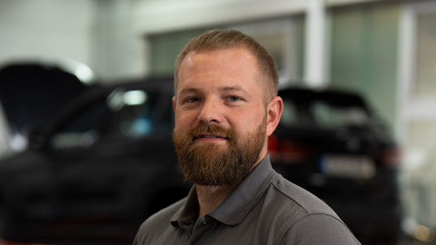 Ein Mann mit Bart in grauem Poloshirt steht vor einem unscharfen schwarzen Auto in einer Werkstatt.