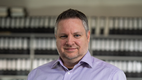 Ein Mann in einem lila Hemd steht vor einem Regal mit Ordnern.