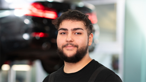 Ein Mann mit dunklem Haar und Bart lächelt vor einem unscharfen Hintergrund mit einem Auto.