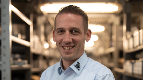 Ein Mann lächelt in einem beleuchteten Lagerraum in die Kamera.