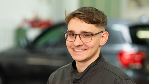 Ein junger Mann mit Brille lächelt in einer Werkstatt, unscharfe Autos im Hintergrund.