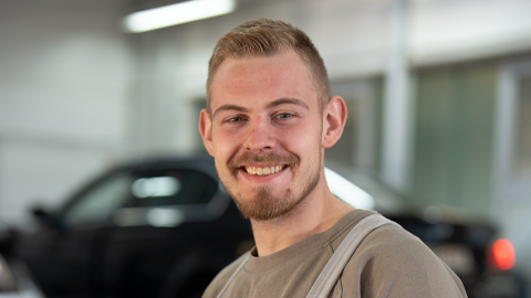 Ein lächelnder Mann vor einem unscharfen Auto in einer Werkstatt.