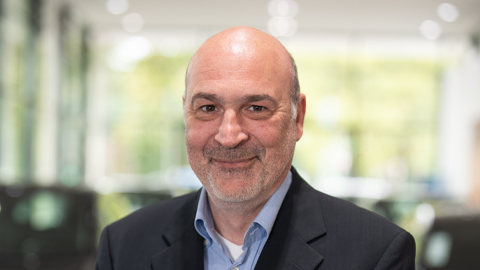 Ein Mann in einem Anzug lächelt in die Kamera; unscharfer Hintergrund mit Fenstern.