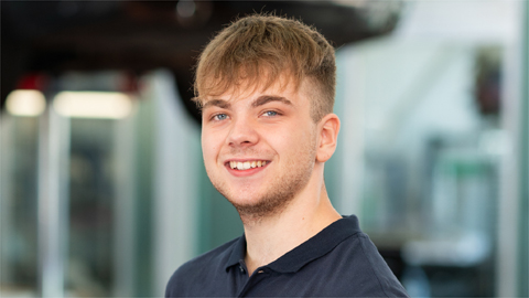 Junger Mann mit dunkelblauem Hemd lächelt in die Kamera, unscharfer Hintergrund mit Werkstattelementen.