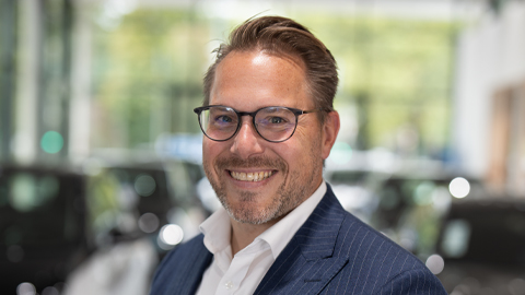 Ein Mann mit Brille lächelt in die Kamera; unscharfer Hintergrund mit Autos in einem Showroom.