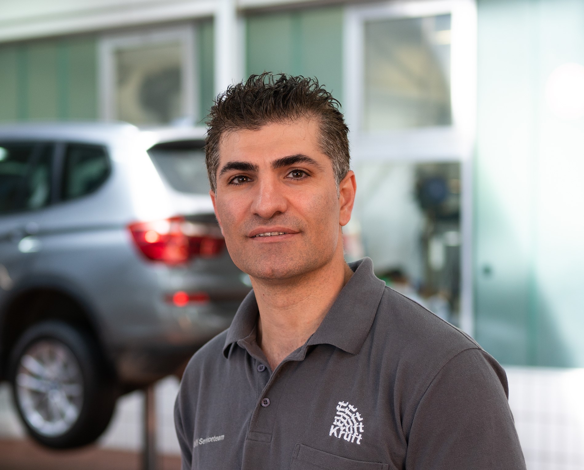 Eine Person in grauem Shirt in Werkstatt, unscharfes Auto im Hintergrund.