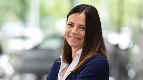 Frau mit dunklem Haar und weißer Bluse lächelt vor unscharfem Hintergrund.