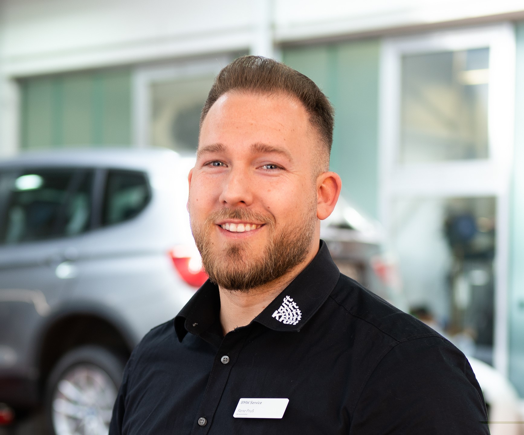 Mann in schwarzem Hemd mit Namensschild in einer Autowerkstatt, unscharfes Auto im Hintergrund.
