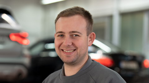 Ein Mann in Werkstattkleidung lächelt, unscharfe Autos im Hintergrund.