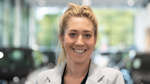 Eine lächelnde Frau in einem hellen Blazer steht in einem Autohaus.