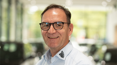 Mann mit Brille lächelt in hellem Raum, unscharfe Autos im Hintergrund.