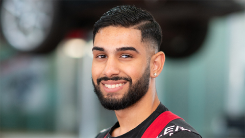 Ein lächelnder Mann in rotem Arbeitsanzug vor unscharfem Hintergrund in einer Werkstatt.