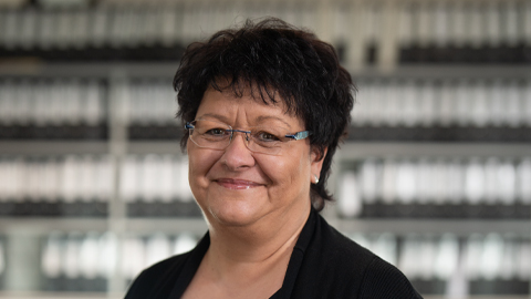 Frau mit Brille und dunklem Haar lächelt vor unscharfem Hintergrund.