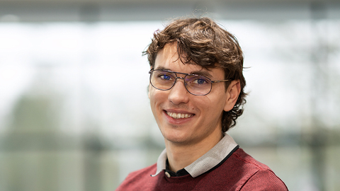 Ein lächelnder Mann mit Brille und braunem Haar vor unscharfem Hintergrund.