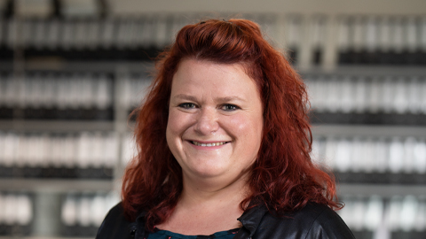 Eine lächelnde Person mit roten Haaren vor einem unscharfen Hintergrund.