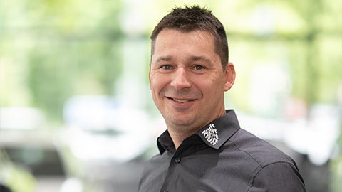 Ein Mann in dunklem Hemd lächelt in die Kamera, Hintergrund unscharf und grünlich.