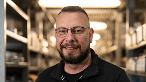 Mann mit Brille und Bart lächelt in die Kamera, Hintergrund unscharf mit Regalen.