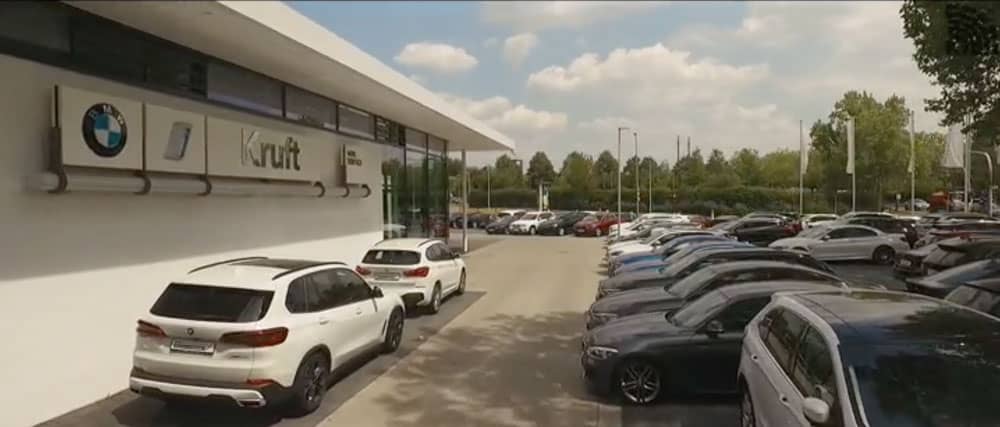 Parkplatz mit vielen Autos vor einem Gebäude mit Kruft-Logo und BMW-Schild, Bäume im Hintergrund.